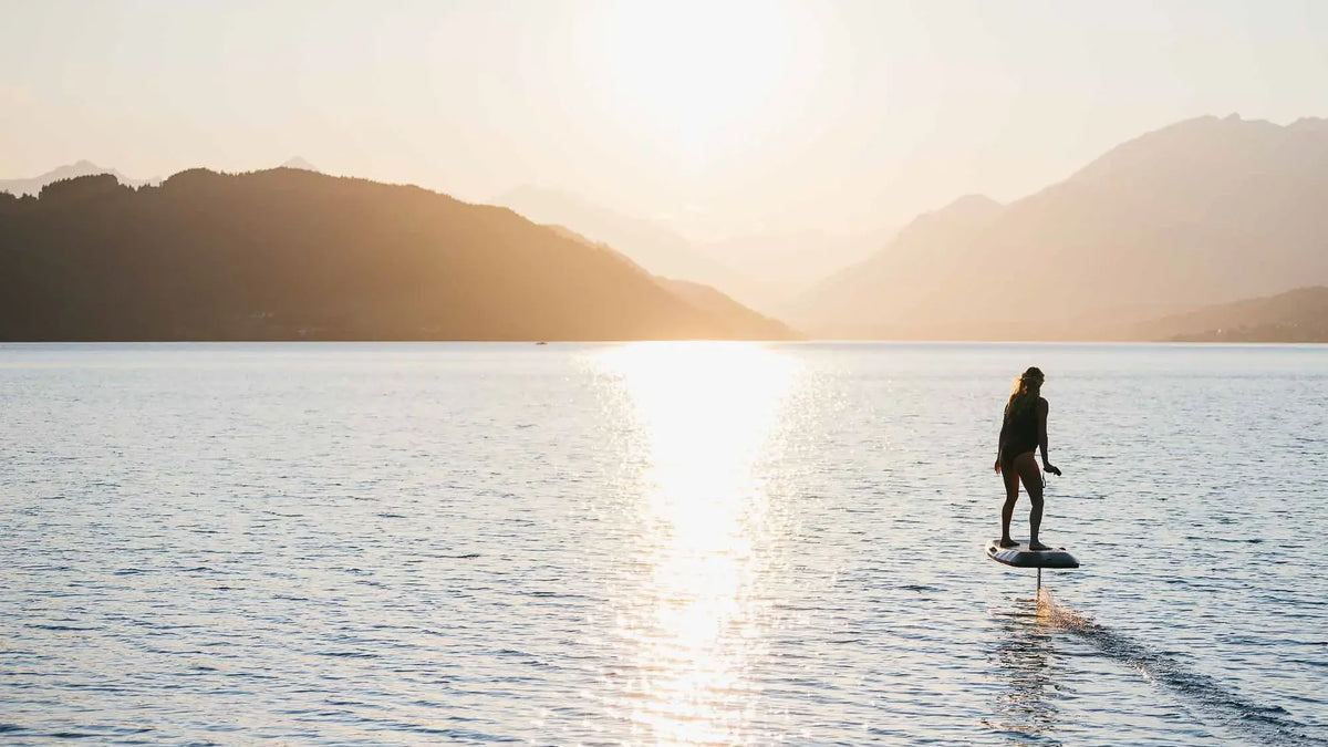 A woman rides an eFoil across a mountain lake at sunset, the board flying just above the water