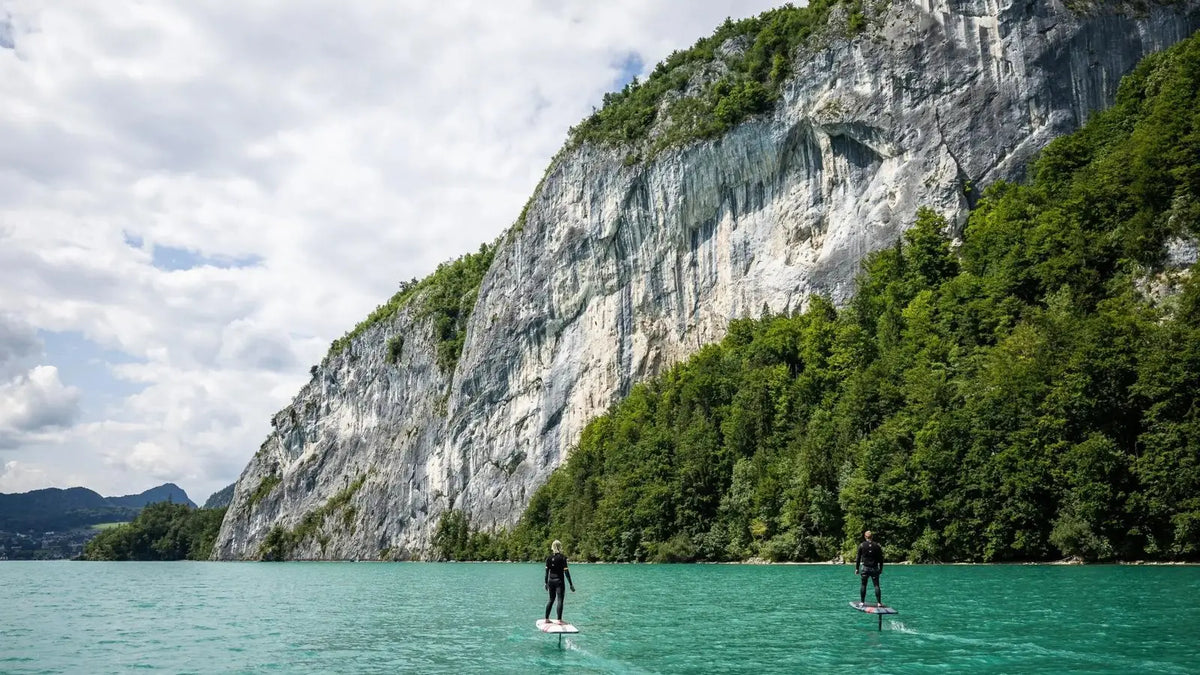 Two riders gliding on eFoils across a turquoise mountain lake below a steep limestone cliff