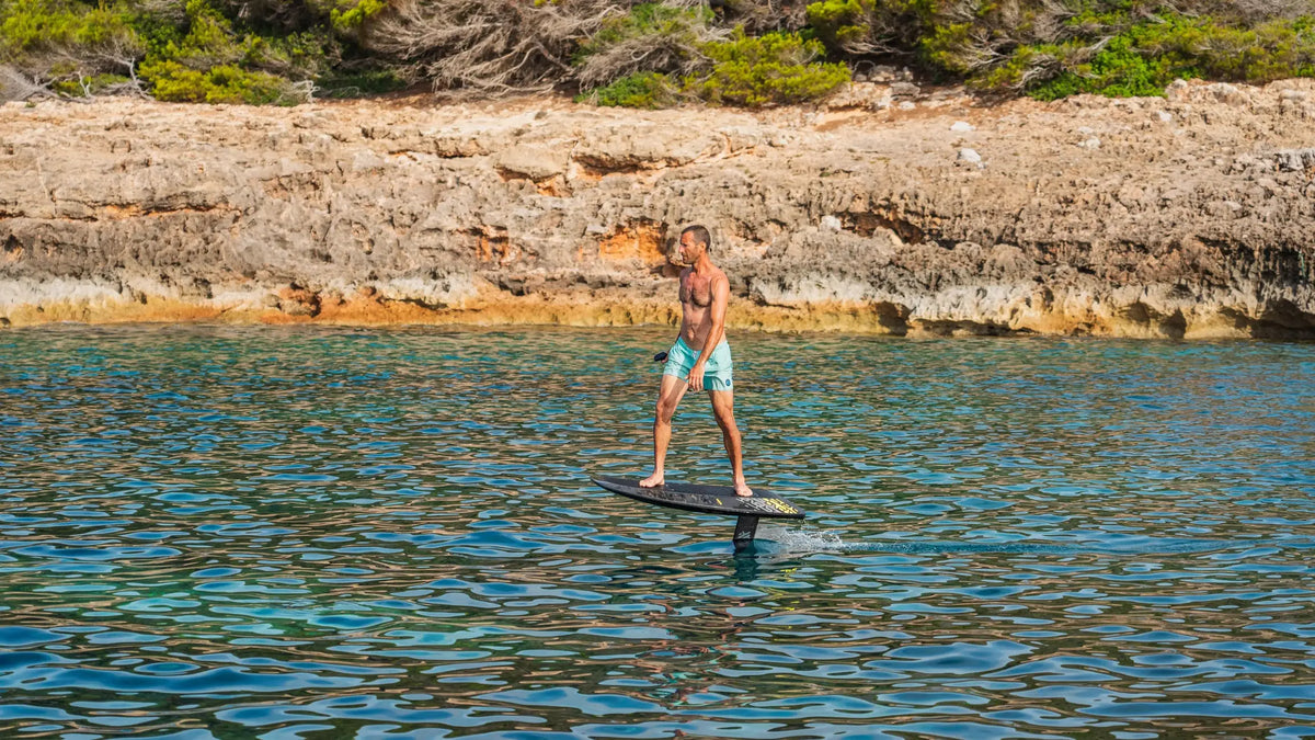 A rider in turquoise shorts flying on an eFoil board above clear water along a rocky coast